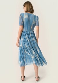 Vestido midi azul en capas con rayas verticales, mangas cortas abullonadas y una textura ligera y fluida. Combinado con sandalias negras de tiras.