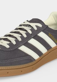 Γκρι σουέτ Adidas αθλητικό με λευκά κορδόνια, τρεις λευκές ρίγες, χρυσή επιγραφή «Spezial» και καφέ λαστιχένια σόλα.
