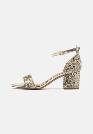 Sandale à talon bloc en paillettes dorées avec une bride métallique à la cheville et un bout ouvert. Doublure intérieure lisse et surface extérieure texturée en paillettes.