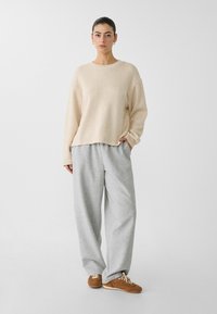 Beige Oversize-Pullover mit Raglanärmeln, kombiniert mit hellgrauen, weit geschnittenen Jogginghosen und braunen Sneakern mit beigefarbenen Schnürsenkeln.