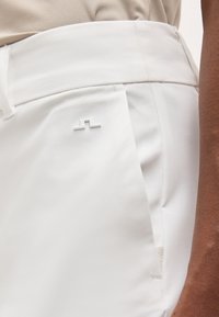 Κοντινή λήψη λευκών ψηλόμεσων παντελονιών με ένα μικρό λευκό λογότυπο στην εμπρόσθια τσέπη, που φοριούνται από άτομο με καφέ επιδερμίδα.