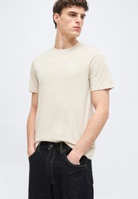 Νεαρός άνδρας που φοράει μπεζ κοντομάνικη μπλούζα και σκούρο τζιν, στέκεται με τα χέρια στις τσέπες, κοιτάζοντας προς τα πλάγια σε απλό φόντο.