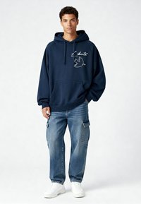 Sweat à capuche bleu marine avec une poche kangourou à l'avant, orné d'une broderie blanche d'oiseau et du mot « liberté ». Associé à un jean cargo bleu clair de style cartoon.