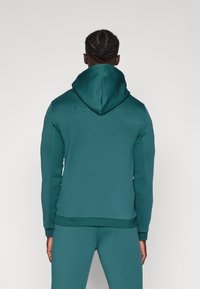 Sweat à capuche en bleu sarcelle, fabriqué en tissu texturé avec des poignets et un ourlet côtelés. Présente un logo en relief sur le haut du dos.
