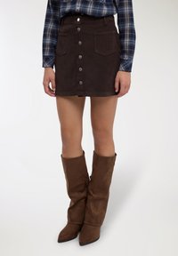 Minigonna marrone in velluto a coste con chiusura frontale a bottoni, tasche applicate e una texture sottile. Abbinata a stivali marroni sfilati fino al ginocchio.