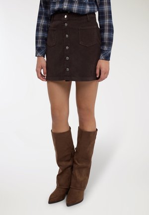 Minigonna marrone in velluto a coste con chiusura frontale a bottoni, tasche applicate e una texture sottile. Abbinata a stivali marroni sfilati fino al ginocchio.