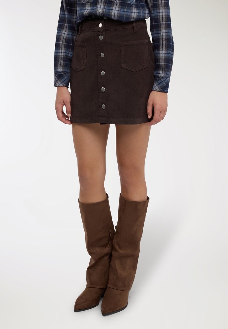 Minigonna marrone in velluto a coste con chiusura frontale a bottoni, tasche applicate e una texture sottile. Abbinata a stivali marroni sfilati fino al ginocchio.