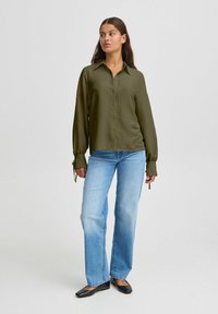 Olive green button-up shirt with long sleeves and tie detail at the cuffs, paired with light blue straight-leg jeans and black flats.