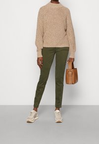 Světle hnědý pletený svetr s vysokým límcem, zelené slim-fit kalhoty, béžové tenisky a malá hnědá kabelka.