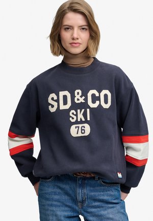 Ung kvinde med kort blondt hår iført en navy sweatshirt med teksten "SD & CO SKI 76" og blå jeans, stående med hænderne i lommerne.