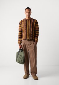 Gebreide trui met een bruin, geel en zwart gestreept patroon; beige wijde broek; groene gewatteerde tas; bruine pantoffels. Neutrale achtergrond.