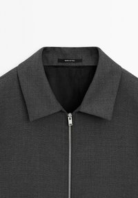 Grå zip-up jakke med flad krave, lavet af tekstureret stof. Indvendig foret med sort, med en metal lynlås og "MADE IN ITALY" label.