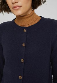 Cárdigan de punto navy con cuello redondo, seis botones de madera, usado sobre un cuello de tortuga de canalé marrón. Textura suave, diseño casual.