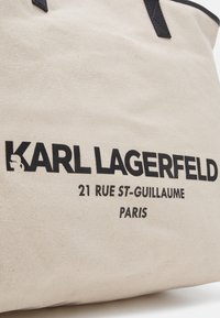Beige canvas tote bag met zwarte tekst. Bevat branding "KARL LAGERFELD" en adres "21 RUE ST-GUILLAUME PARIS." Zwarte lederen accenten.