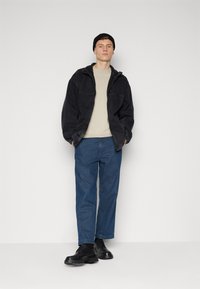 Černá manšestráková bunda na zip, béžové tričko s kulatým výstřihem, modré volné džínsy a černé boty dotvářejí outfit. Postavte se na neutrální pozadí.