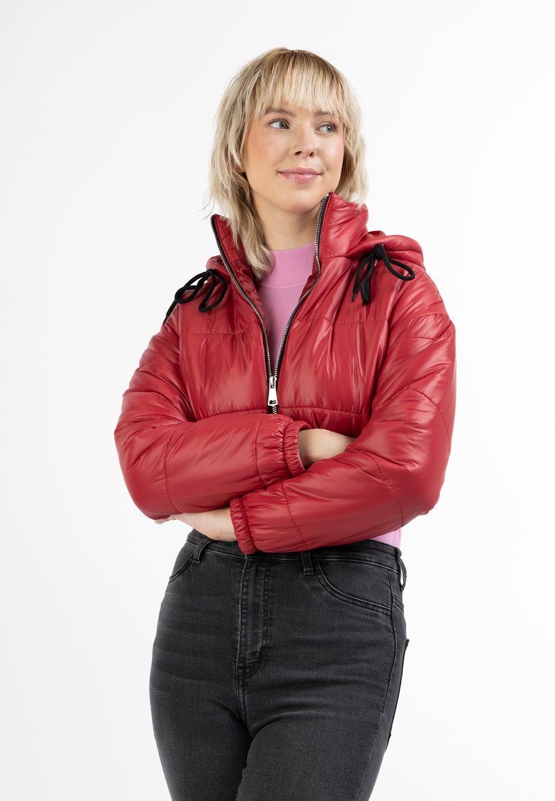 Giacca a vento rossa con finitura lucida, apertura con zip e lacci neri sul cappuccio. Indossata sopra una maglietta rosa e jeans neri a vita alta.