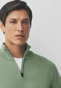 Jonge man met kort donker haar en blauwe ogen die een groene half-rits trui over een wit shirt draagt, tegen een effen lichte achtergrond.