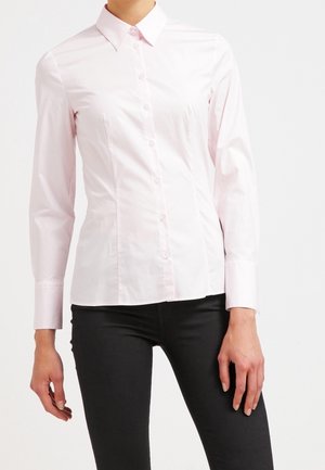 Personne portant une chemise rose clair ajustée à manches longues boutonnée et un pantalon noir, debout devant un fond blanc uni.