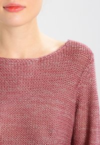 Pull en maille dans une teinte rose poussiéreux avec un décolleté large et décontracté. Il présente une finition texturée avec des fils métalliques subtils, offrant un aspect doux.