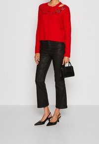 Cardigan court rouge à fermeture boutonnée, orné de sequins sur l'épaule ; associé à un pantalon noir évasé textur�é et des talons pointus.