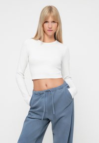 Bijeli, dugih rukava crop top s rebrastom teksturom uparen s plavim duksericama koje imaju podesivi pojas i bočne džepove.