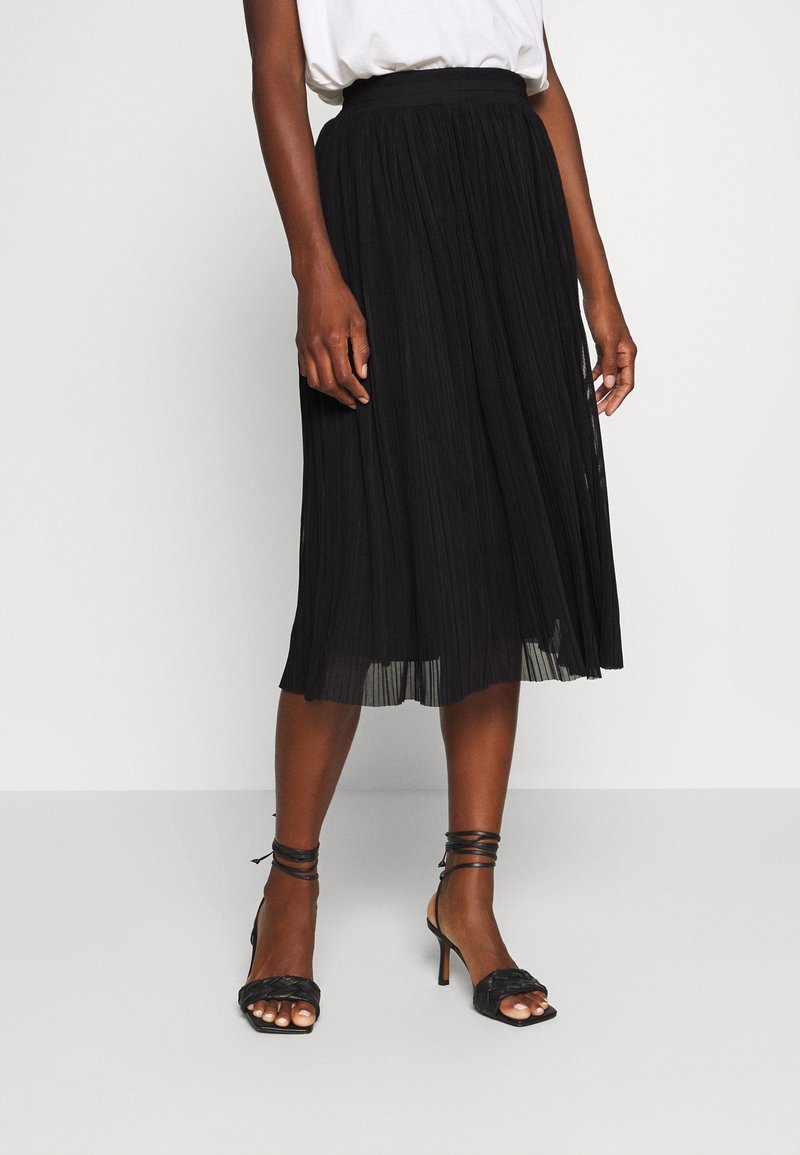 Femme portant une jupe midi plissée noire, un haut blanc et des sandales à talons hauts noires à brides, debout sur un sol clair et uni.