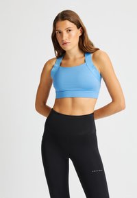 Sujeción deportiva de color azul claro con tirantes anchos, hecha de una tela elástica. Leggings negros de cintura alta con un sutil logotipo en el costado.