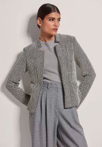 Schwarz-weißer, gemusterter Blazer mit strukturiertem Design und Reverskragen, kombiniert mit einem grauen Sweater und hochgeschnittenen grauen Hosen.
