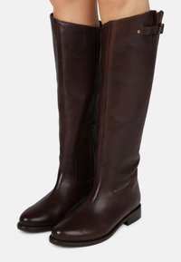 Botas de couro castanho até ao joelho, com uma textura suave, bico arredondado, fecho lateral e um pequeno detalhe em fivela na parte superior. Sola plana.