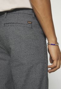 Gros plan d'une personne portant un pantalon gris texturé avec l'étiquette « BOSS » sur la poche arrière et un bracelet argenté avec des perles bleues au poignet.