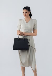 Borsa tote nera con texture, manici doppi, forma rettangolare e dettaglio del marchio dorato, tenuta da una persona in un abito avvolgente beige chiaro.