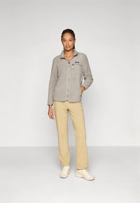 Hall halli fliisist jakk esiga lukustuse, kõrge krae ja rinnataskuga. Kombineeritud beežide pükste ja heledate jalatsitega.