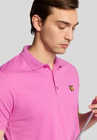 Pink polo shirt in a breathable fabric, featuring a two-button placket and a black embroidered logo on the left chest, smooth texture.