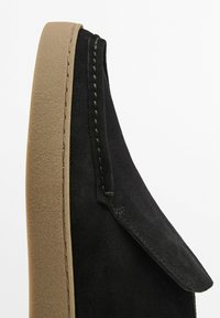 Černá semišová bota s kulatou špičkou a taupe podrážkou. Obsahuje kontrastní švy podél okraje a skládací design klapky.