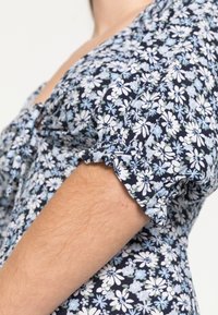 Květinové šaty s námořnickým podkladem, zdobené bílými a světle modrými květinami. Krátké nabírané rukávy s volánovým lemem a detail výstřihu ve tvaru klíčové dírky.