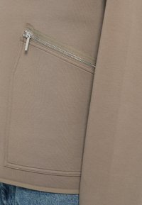 Sudadera beige con una tela texturizada que presenta un bolsillo con cremallera en el costado. El bolsillo tiene una cremallera plateada y detalles de costura limpios.