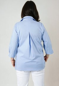 Chemise bleu clair à boutons avec un pli au dos, manches longues retroussées aux poignets, et col. Le tissu semble lisse et léger.