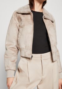 Krótkie beżowe kurtka z miękkiego sztucznego futra z kołnierzem, przednim zamkiem błyskawicznym i ściągaczami na rękawach, w połączeniu z beżowymi spodniami z wysokim stanem.