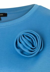 Blauwe stoffen top met een ronde halslijn en een driedimensionale blauwe stoffen rozenaccent aan de linkerkant. Glad textuur.