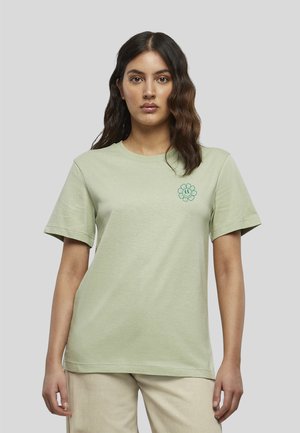 Jeune femme aux cheveux foncés et ondulés porte un t-shirt vert clair avec un petit motif de fleur souriante sur la poitrine gauche, sur un fond uni.