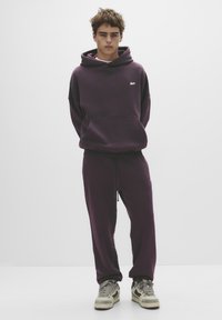Hanorac violet cu glugă mare și buzunar frontal, asortat cu pantaloni tapered. Caracteristici: cordon la talie și detaliu cu logo. Adidași vizibili.