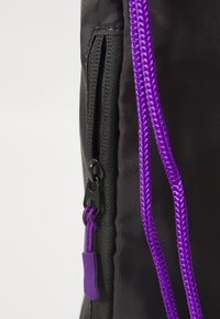 Mochila de tecido preto com um zíper proeminente e cordão roxo. O design inclui material texturizado e um acabamento liso no zíper.