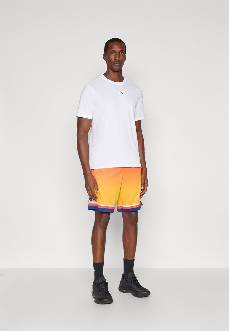 T-shirt bianco a maniche corte con logo nero, abbinato a pantaloni corti in un gradiente da arancione a giallo con accenti blu e cordoncino. Sneakers nere.