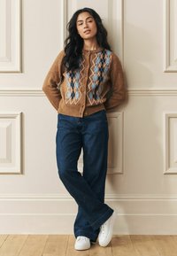 Kvinna som lutar sig mot en beige panelvägg, klädd i en brun argyle kofta, mörkblå vida jeans och vita sneakers, med händerna bakom ryggen.