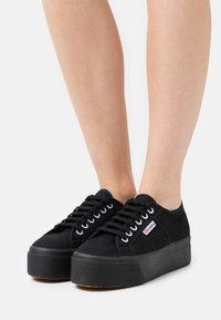 Sorte kanvas sneakers med teksturert overdel, hevet plattformsåle og hvite øyer, med et lite merke på siden.