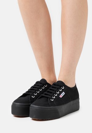 Schwarze Leinenschuhe mit strukturiertem Obermaterial, erhöhtem Plateausohle und weißen Ösen, mit einem kleinen Markenetikett an der Seite.