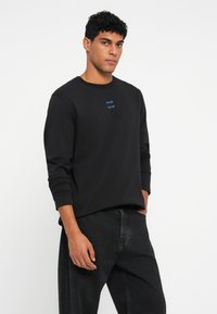 Jauns vīrietis ar viļņainiem matiem, ģērbies melnā garajās piedurknēs krekls ar maziem ziliem uzrakstiem un melnās džinsās, stāv ar vienu roku kabatā.