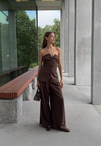 Vrouw in een bruine top met naadloze strepen zonder bandjes en een broek met wijde pijpen, met een kleine handtas, staand bij betonnen zuilen en een houten bankje buiten.