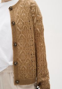 Lichtbruine gebreide cardigan met een opengewerkt geometrisch patroon, ronde halslijn en grote donkere knopen aan de voorkant.