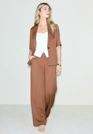 Žena s dlhými blond vlasmi, oblečená v hnedých širokých nohaviciach a krátkom blejzri s krátkym rukávom, bielom zapínanom topu, kráča bosá po svetlej podlahe.